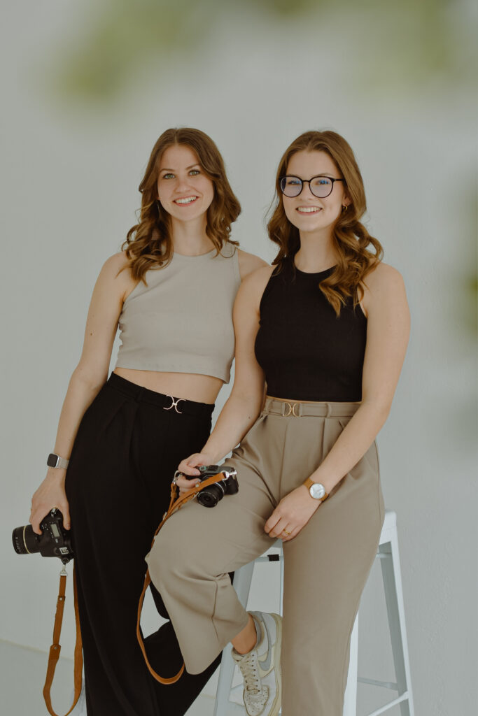 Annika & Alisa - Fotografinnen-Duo Zwei Fotografinnen stehen nebeneinander und lächeln in die Kamera. Sie halten jeweils eine Kamera in der Hand, im Hintergrund ist ein natürliches Setting zu sehen.